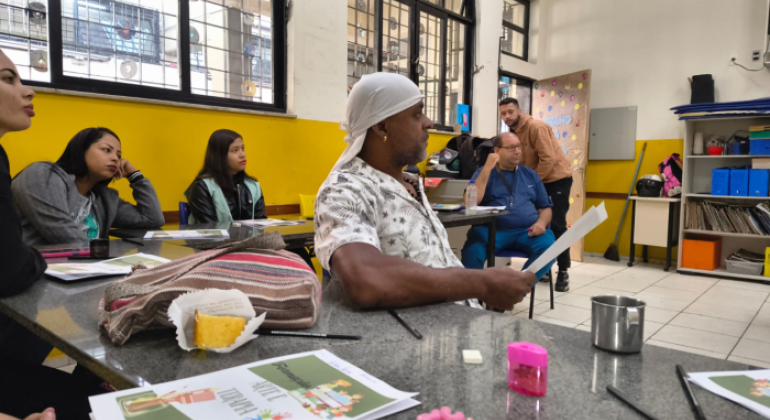 #paratodosverem: em uma sala de aula com paredes amarelas e janelas gradeadas, um grupo de pessoas adultas está sentado ao redor de mesas, prestando atenção a alguém fora de cena. Sobre a mesa, há panfletos coloridos com o título “Arte e terapia”, lápis, apontador, uma caneca metálica e um pedaço de bolo em um guardanapo. Ao fundo, um homem de blusa marrom está em pé.