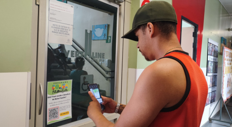 #paratodosverem: em um corredor, um homem com boné e camiseta vermelha e preta lê o QR Code em um panfleto colado em uma porta; ele está virado de lado para a câmera, com seu celular apontado para o panfleto.