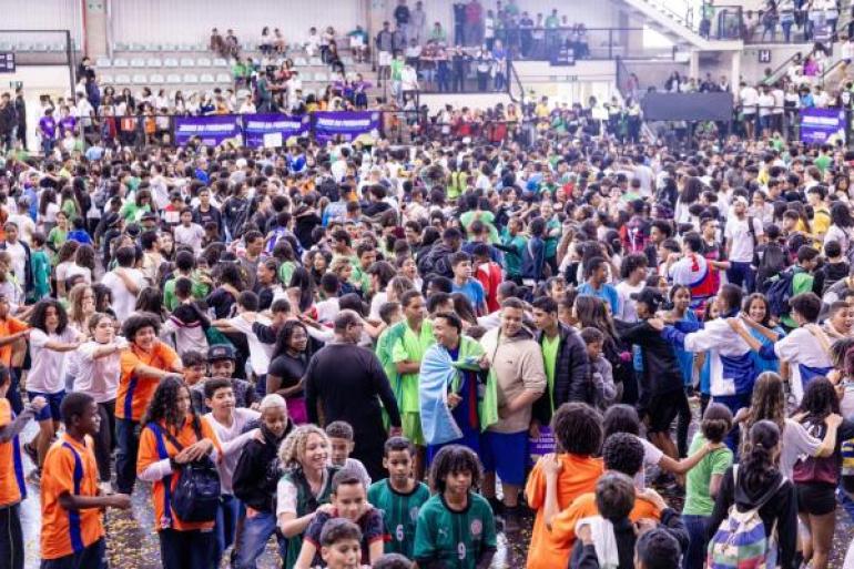#paratodosverem: Uma multidão de estudantes, jovens e adolescentes, de camisetas coloridas, celebra a abertura dos Jogos da Primavera em uma quadra. Há uma atmosfera de energia e alegria, com muitas pessoas sorrindo e interagindo. Confetes estão espalhados pelo chão. A cena transmite a sensação de um evento escolar vibrante.