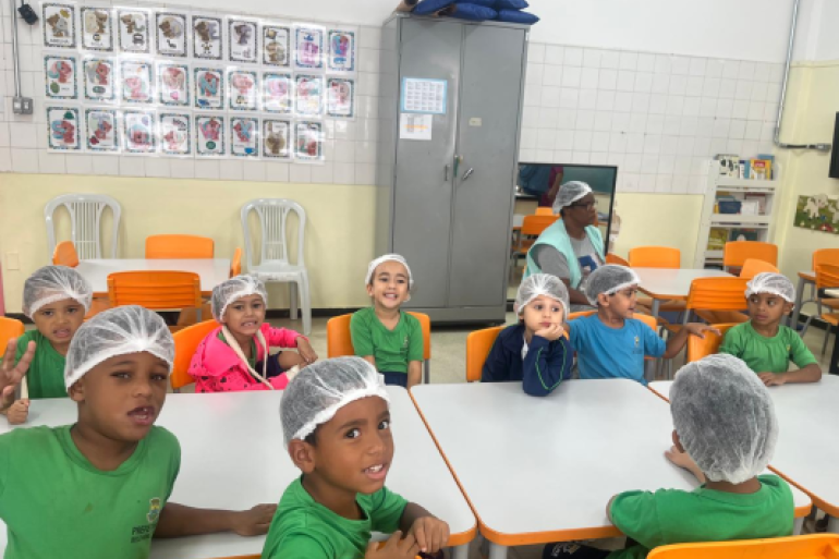 #paratodosverem: Em uma sala de aula clara, com mesas e cadeiras laranja e brancas, um grupo de crianças está sentado em torno de duas mesas. Todas usam toucas brancas de cabelo e camisetas de uniforme. Algumas sorriem para a câmera, exibindo entusiasmo em um momento de preparo para uma atividade.