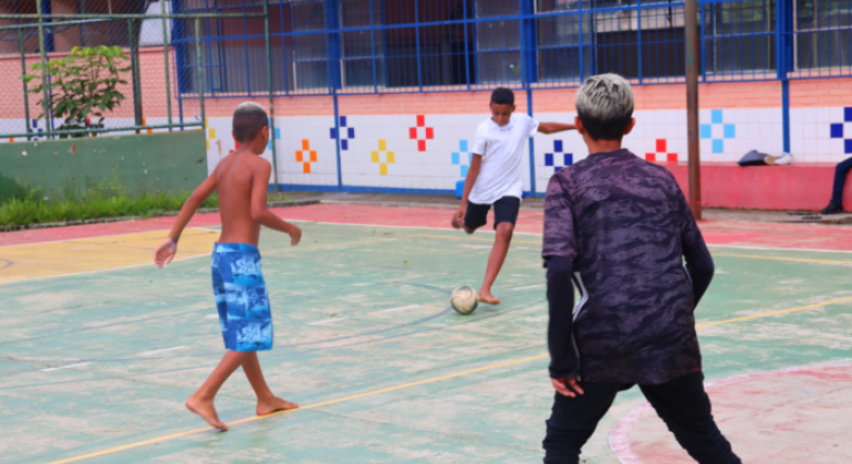 #paratodosverem: Fotografia colorida de três crianças jogando futebol na quadra da escola. Duas crianças estão de costas para a câmera e uma está de frente, chutando a bola.