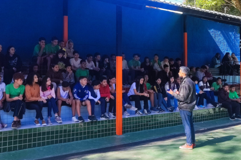 Estudantes fazem curso de customização de camisas para aprender sobre a cultura do hip hop #paratodosverem: A imagem mostra um grupo grande de estudantes sentados em arquibancada coberta, dividida em dois níveis, com paredes azuis e colunas laranja. Eles observam atentamente um homem de jaqueta preta e calça jeans que está em pé na frente, gesticulando enquanto fala. O espaço é iluminado pela luz natural e tem piso verde.