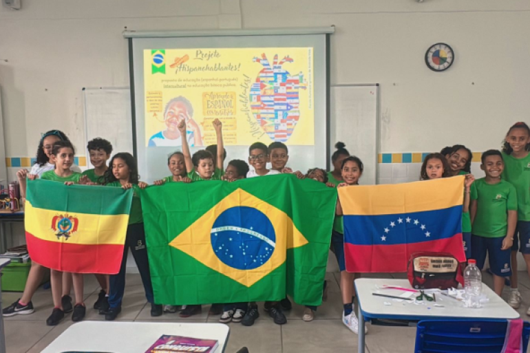 O objetivo é integrar estudantes brasileiros e migrantes #paratodosverem: Em uma sala de aula, um grupo de crianças sorridentes segura três bandeiras: Bolívia, Brasil e Venezuela. No telão ao fundo, o tema do "Projeto Hispanohablantes" é exibido com um mapa de corações