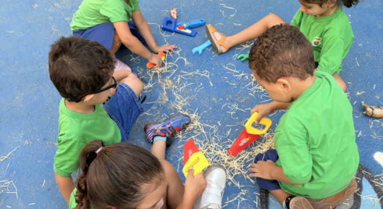 #paratodosverem: Cinco crianças sentadas no chão azul participam de uma atividade. Elas usam uniformes verdes e exploram ferramentas de brinquedo enquanto manuseiam palhas espalhadas pelo chão. Uma menina de cabelo preso em rabo de cavalo baixo e cacheado observa, enquanto outras, com cabelos soltos ou presos, interagem em grupo, formando uma roda.