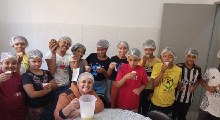 Aprendizado com sabor: crianças exploram a alimentação de forma divertida. #paratodosverem: Na imagem, um grupo de crianças posa sorridente em uma cozinha, todas usando toucas de proteção. Elas seguram copinhos com suco e demonstram alegria pela atividade. Uma mulher, também de touca, está sentada à frente, segurando uma jarra com a bebida, enquanto uma das crianças exibe um abacaxi. A cena transmite aprendizado e diversão em um momento coletivo.