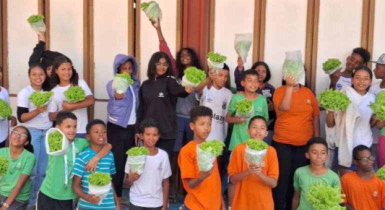 #paratodosverem: Um grupo de estudantes posa sorridente segurando pés de alface colhidos na horta escolar. Alguns levantam as hortaliças acima da cabeça, outros exibem à frente do corpo. Vestindo uniformes em verde, branco, laranja e azul, eles se organizam em frente a uma parede clara com janelas de madeira.