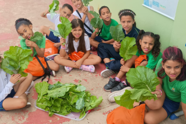 Estudantes aprendem sobre a natureza e alimentação saudável #paratodosverem: A imagem mostra um grupo de crianças sentadas no chão em círculo, sorrindo e segurando grandes folhas verdes de hortaliças. No centro, há uma bandeja cheia de folhas semelhantes. Elas vestem uniformes escolares em azul, branco e verde, e algumas carregam sacolas laranjas.
