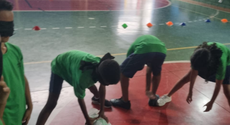 #paratodosverem: Na foto, um grupo de crianças participa de uma brincadeira no ginásio da escola. Usando vendas nos olhos e camisetas verdes, elas se abaixam tentando encontrar objetos no chão, guiando-se apenas pelo tato e pelas orientações dos colegas. A atividade promove cooperação, confiança e diversão em um ambiente educativo e acolhedor.