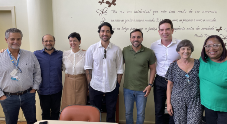 #paratodosverem: a foto mostra um grupo de oito pessoas reunidas em um ambiente interno, posando sorridentes para a câmera. Ao fundo, há uma parede clara decorada com o desenho de algumas borboletas e uma frase do educador Paulo Freire: "Eu sou um intelectual que não tem medo de ser amoroso. Eu amo as gentes e amo o mundo. E é porque amo as pessoas e amo o mundo, que eu brigo para que a justiça social se implante antes da caridade." Na frente do grupo, há uma mesa com cadernos abertos, um copo e um celular.