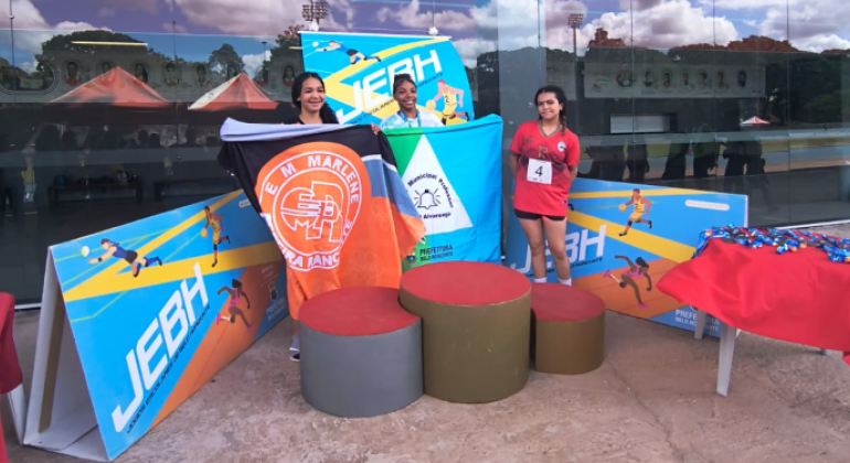 #paratodosverem: a imagem mostra uma cerimônia de premiação dos Jogos Escolares, realizada em um ambiente ao ar livre, em frente a um prédio com fachada de vidro espelhado. Três jovens atletas estão posicionadas sobre um pódio. As atletas que conquistaram o primeiro e o segundo lugar seguram as bandeiras de suas escolas: Professor Daniel Alvarenga e Marlene Pereira Rancante, respectivamente.