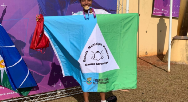 #paratodosverem: Uma menina está em um pódio vermelho segurando uma bandeira com as cores azul, branca e verde. Na bandeira está escrito “Escola Municipal Professor Daniel Alvarenga” e “Prefeitura Belo Horizonte”, além de um sino desenhado. Ela morde uma medalha dourada pendurada no pescoço e segura uma bolsa vermelha.