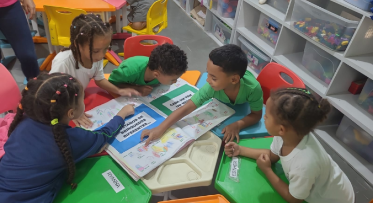 #paratodosverem: na foto, crianças uniformizadas estão reunidas ao redor de uma mesa colorida, interagindo com um livro coletivo cheio de desenhos e frases. As mesas e cadeiras são em tons de verde, vermelho, amarelo e azul. Ao fundo, prateleiras brancas organizadas guardam brinquedos e materiais, que estão dentro de caixas transparentes.