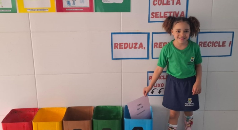 paratodosverem: na foto, uma criança uniformizada sorri ao lado de lixeiras coloridas de coleta seletiva, identificadas como “plástico”, “metal”, “orgânico”, “vidro” e “papel”. Ela deposita uma folha na lixeira azul, destinada ao papel. Ao fundo, cartazes educativos reforçam a importância da reciclagem com os dizeres "Reduza, Reuse, Recicle!"