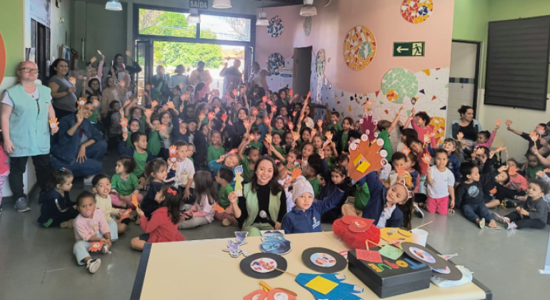 #paratodosverem: na foto, um grande grupo de crianças está reunido em um salão escolar, acompanhado por educadores(as). Muitas delas seguram palitinhos com mãos coloridas de papel. No ambiente, há decorações coloridas nas paredes, com mosaicos e placas de sinalização. À frente, uma mesa exibe discos de papel e materiais de artesanato.