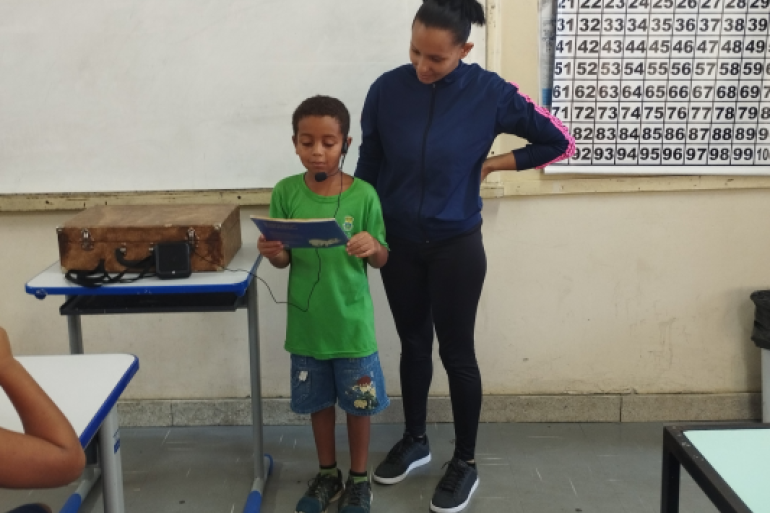 #paratodosverem: Um menino de uniforme verde e bermuda jeans lê um livro em voz alta, apoiado por um microfone de cabeça. Ele está no centro de uma sala de aula e, ao lado dele, uma professora o observa com carinho. A cena mostra um momento de leitura e apresentação oral em frente a um quadro branco e um cartaz numérico.