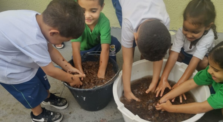 #paratodosverem: Seis crianças pequenas estão em um pátio, explorando dois baldes grandes com terra e água. Elas estão de pé ou agachadas, com as mãos imersas nos baldes, sorrindo e interagindo entre si. Algumas usam camisetas verdes e outras brancas, com shorts azuis. Ao fundo, há uma parede amarela.