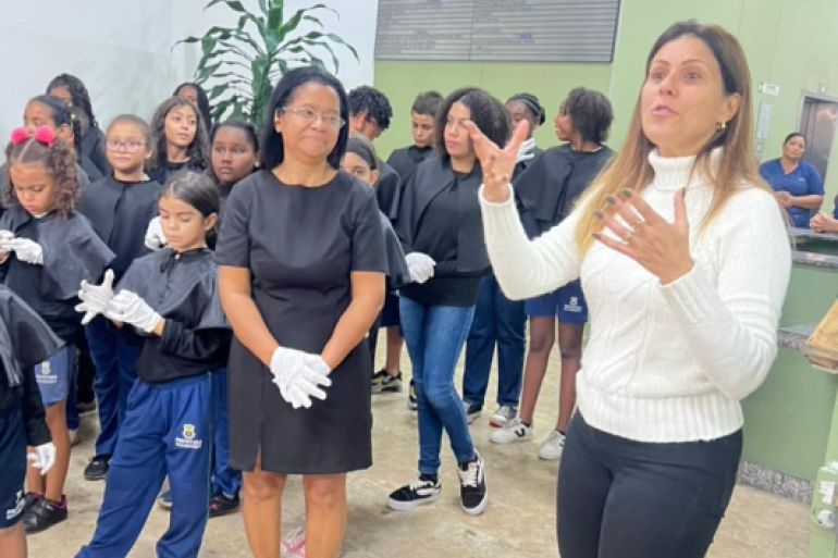 Os estudantes participam de vários projetos. #paratodosverem: na imagem, ao centro, uma mulher de vestido preto também usa luvas, e ao lado dela uma mulher de blusa branca de gola alta gesticula enquanto fala. Ao fundo, um grupo de crianças e adolescentes está reunido. Eles vestem uniforme azul com sobreposição preta e usam luvas brancas