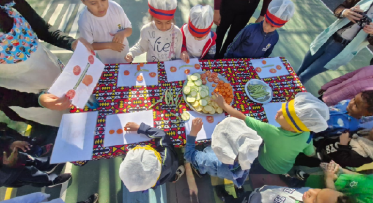 #paratodosverem: Crianças com toucas de cozinha estão em volta de uma mesa colorida ao ar livre. Sobre a mesa há pratos com rodelas de tomate, pepino, abobrinha e cenoura. Algumas crianças mexem nos legumes e outras observam figuras em folhas de papel.