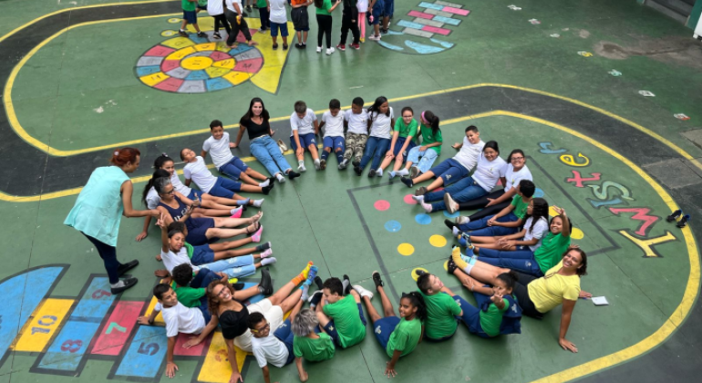 #paratodosverem: A imagem mostra um grupo de crianças e adolescentes sentados(as) no chão de um pátio escolar. Eles(as) estão vestindo uniformes escolares, predominantemente nas cores azul e verde, e alguns(mas) estão usando meias coloridas e descombinadas, criando um visual divertido e chamativo. Algumas pessoas adultas também estão presentes, ajudando na atividade. O chão do pátio tem desenhos coloridos de jogos tradicionais, como amarelinha e twister.