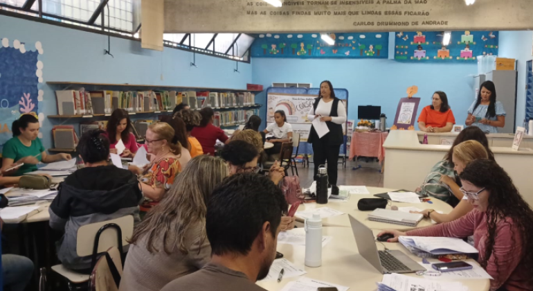 #paratodosverem: na foto, em uma biblioteca escolar, vários(as) educadores(as) estão sentados(as) em mesas redondas, concentrados(as) em leituras e anotações. Ao fundo, uma mulher conduz a apresentação em pé, segurando papéis. Atrás dela, há uma mesa com garrafa de café e um lanche. Outras pessoas estão próximas ao balcão da biblioteca. À esquerda, há estantes repletas de livros. 