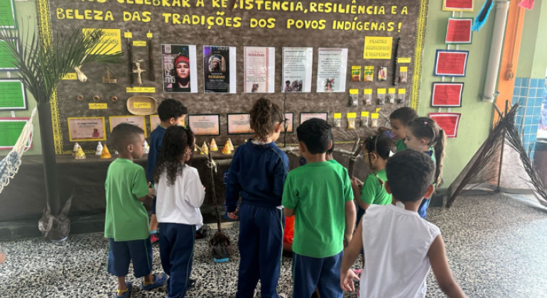 #paratodosverem: na imagem, crianças uniformizadas observam com atenção um mural escolar com o cartaz: "Vamos celebrar a resistência, resiliência e a beleza das tradições dos povos indígenas!". O painel conta com cartazes informativos, objetos artesanais e elementos decorativos, como penas e palha, criando um ambiente de aprendizado visual e interativo.
