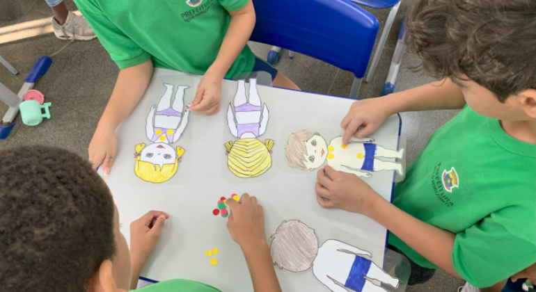 #paratodosverem: na imagem, quatro crianças, com camisetas verdes do uniforme escolar, estão reunidas em torno de uma mesa branca. Sobre ela, há bonecos(as) de papel e círculos adesivos coloridos, representando o “Semáforo dos Toques". As crianças colam os adesivos nos(as) bonecos(as). Ao fundo, há cadeiras azuis e parte do ambiente escolar.