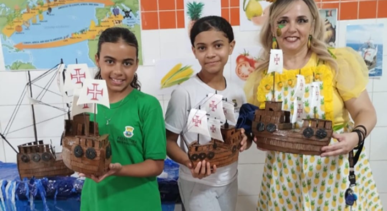 #paratodosverem: a imagem mostra três pessoas em destaque: duas alunas e uma professora, todas segurando maquetes de caravelas portuguesas feitas com material reciclável. As alunas usam uniformes (uma verde, outra branco) e a professora veste blusa amarela, avental com estampa de abacaxi e enfeite de abacaxi na cabeça. Ao fundo, há uma parede de azulejos brancos e alaranjados, com um mapa e uma maquete do mar com uma caravela.