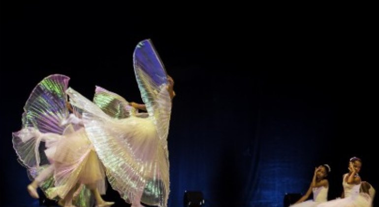 #paratodosverem: na imagem, seis bailarinas estão em um palco. Quatro dançam ao centro com grandes leques iluminados, que lembram asas de borboleta, criando um efeito etéreo. Duas outras estão sentadas à direita, observando encantadas. O fundo é escuro, destacando as luzes e o movimento dos véus brilhantes.