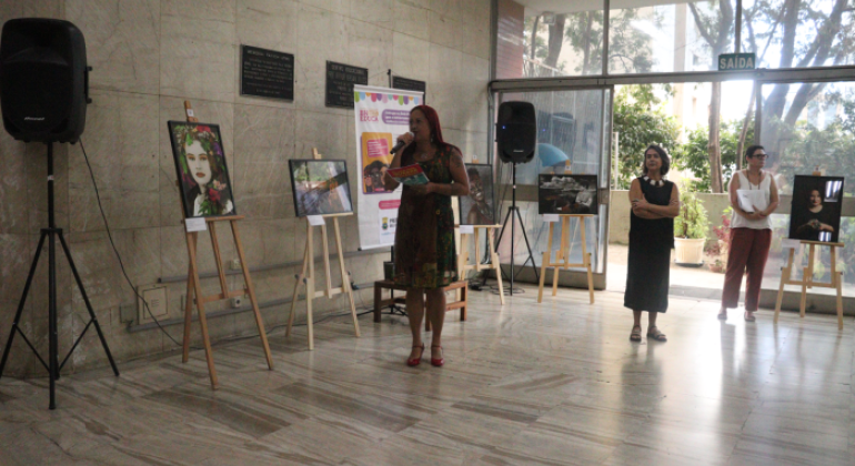 #paratodosverem: A imagem mostra a entrada da Smed, onde uma mulher segura um microfone e um panfleto enquanto discursa, cercada por obras fotográficas expostas em cavaletes. Duas outras mulheres acompanham a apresentação, uma de braços cruzados e outra segurando papéis. O ambiente conta com grandes portas de vidro que permitem a entrada de luz natural. Ao fundo, aparecem outras plantas em vasos e algumas árvores.