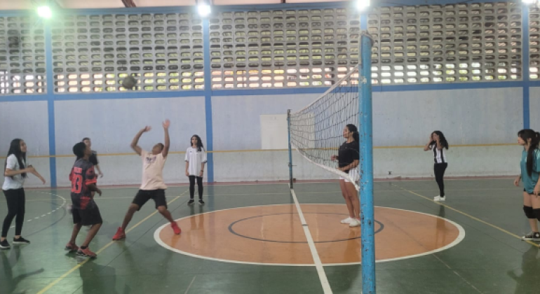 #paratodosverem: em uma quadra poliesportiva, 9 alunos jogam voleibol. À esquerda da rede, é possível ver 5 jovens e à direita, 4. Um dos jovens à esquerda está com as mãos levantadas para receber e jogar a bola que está no ar, em sua direção.