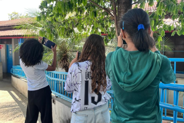 #paratodosverem: Três meninas estão de costas, cada uma segurando um celular para tirar fotos de uma árvore com folhas verdes. Elas estão em área externa, próxima a um prédio com telhado vermelho e muros azuis, cercada por grades azuis. Uma veste camiseta branca, outra camiseta clara com número 13 e a terceira, um casaco verde.