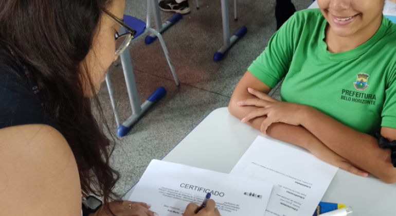 #paratodosverem: A imagem mostra uma sala de aula, com duas pessoas em destaque. À esquerda, uma mulher de cabelos longos e escuros, usando óculos, está assinando um certificado sobre uma mesa. À direita, uma jovem estudante, vestindo uniforme verde, sorri com os braços cruzados sobre a mesa. Ao lado, é possível ver outras cadeiras e carteiras escolares. 