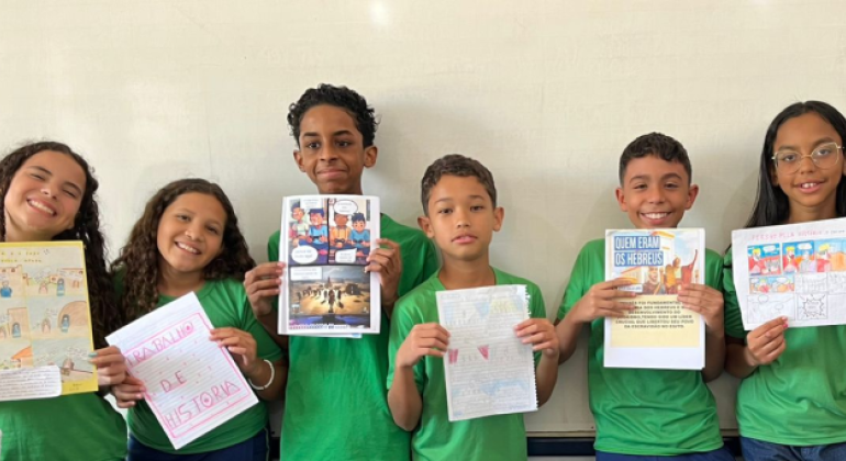 #paratodosverem: na imagem, um grupo de crianças posa sorridente em frente a um quadro branco, exibindo trabalhos de História. Elas estão em pé e vestem uniforme escolar e seguram produções coloridas com textos, desenhos e histórias em quadrinhos sobre povos antigos. 