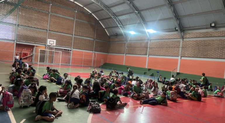 Na quadra de uma escola, seis filas de crianças sentadas com mochilas ao lado aguardam a saída, interagindo entre si. O espaço tem chão colorido, arquibancada verde, parede laranja, tabela de basquete, trave com rede e claraboias no teto que iluminam o ambiente.