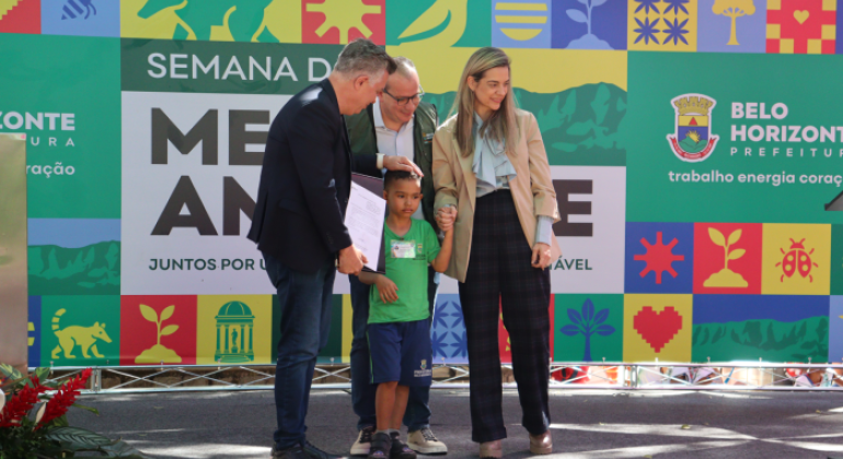 #paratodosverem: na imagem, o Prefeito e os(as) secretários(as), trajados(as) formalmente, posam junto a um aluno da RME-BH para uma foto, em primeiro plano. Ao fundo, há um banner, referente à Semana do Meio Ambiente, contendo elementos coloridos que representam a fauna e a flora.