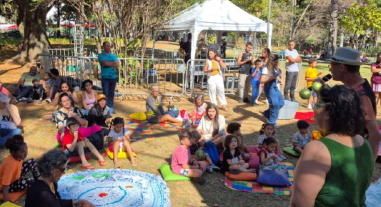#paratodosverem: em um parque, crianças e adultos estão sentados em almofadas e toalhas coloridas no chão, atentos a uma atividade cultural. Duas pessoas em pé, uma com chapéu e microfone, conduzem a apresentação. Ao fundo, árvores, barracas e pessoas circulando compõem o ambiente ao ar livre.