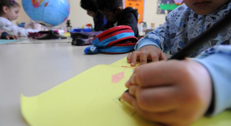 #paratodosverem: a foto mostra crianças em uma sala de aula envolvidas em atividades escolares. Em primeiro plano, uma criança segura um lápis preto e desenha em uma folha amarela, com um traço visível em vermelho. Ela veste uma blusa azul com estampas. Ao fundo, outras crianças estão concentradas em suas tarefas, e, sobre a mesa, há materiais escolares, como estojo e um globo terrestre.