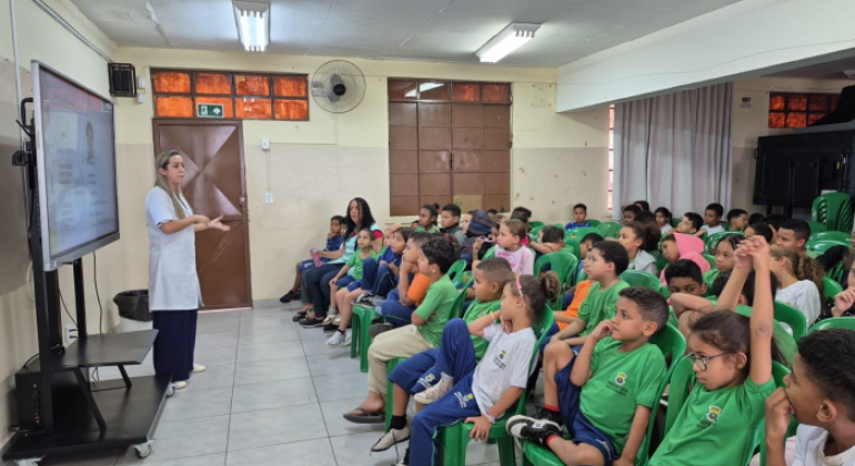 #paratodosverem: sala de aula com fileiras de cadeiras verdes ocupadas por crianças em uniforme escolar, atentas a uma apresentação. À frente, uma mulher de jaleco branco fala e gesticula, ao lado de uma tela digital ligada. O espaço tem paredes claras, janelas com vidros alaranjados e cortina cinza ao fundo.