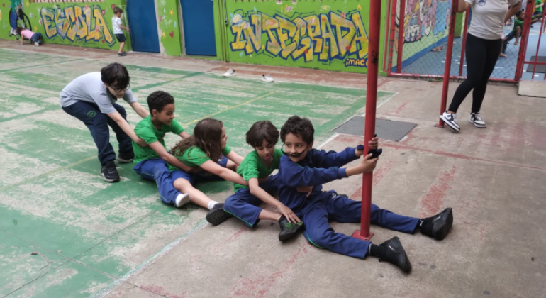 #paratodosverem: em uma quadra escolar com paredes grafitadas, cinco crianças, com uniformes verdes, azuis e branco, participam de uma brincadeira em fila, sentadas no chão e segurando umas nas outras. O primeiro da fila, com um óculos de sol fora do lugar e roupa azul, segura firme em um poste vermelho. Ao fundo, outras crianças brincam e uma educadora observa a cena sorridente.