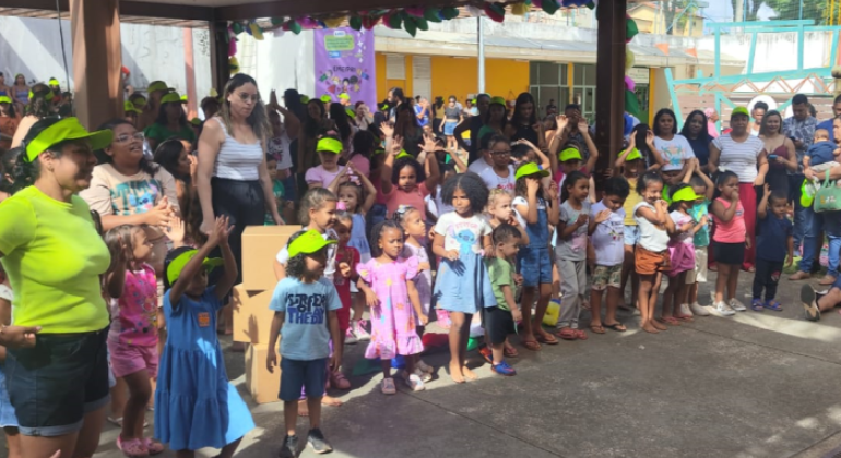 #paratodosverem: a foto mostra uma confraternização com crianças pequenas e pessoas adultas reunidas em um momento coletivo de evento comemorativo ao ar livre. As crianças, muitas usando viseiras verdes, acompanham uma apresentação. 