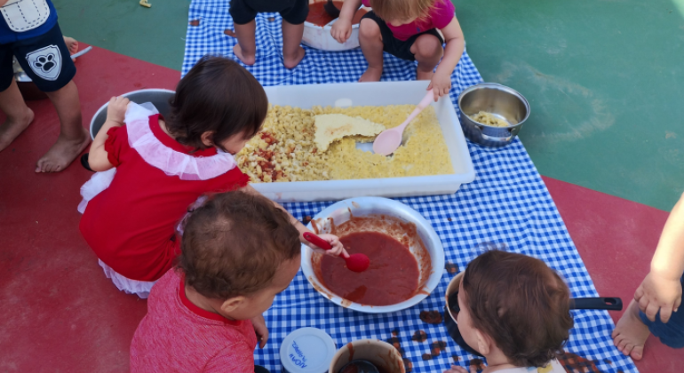 #paratodosverem: crianças pequenas brincam sentadas no chão, sobre toalha xadrez branco com azul. Elas manipulam bandejas com macarrão cozido e tigelas com molho vermelho, usando colheres e as mãos. A atividade acontece ao ar livre, em piso colorido de verde e vermelho. Algumas crianças estão descalças e vestidas com roupas leves e coloridas.