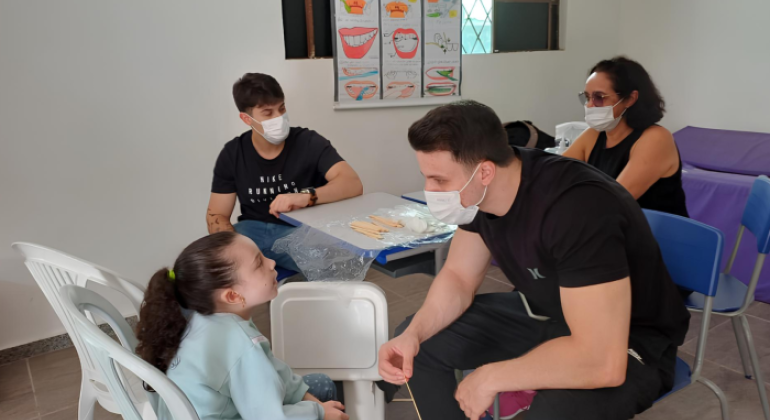 #paratodosverem: em uma sala de paredes brancas, um jovem de camiseta preta e máscara conversa com uma estudante de cabelo preso e blusa azul, que está sentada em frente a ele, que segura um palito de madeira. Ao fundo, estão sentados(as) outro jovem e uma mulher, ambos de máscara. Na parede, há um cartaz pendurado na janela, ilustrando cuidados bucais.
