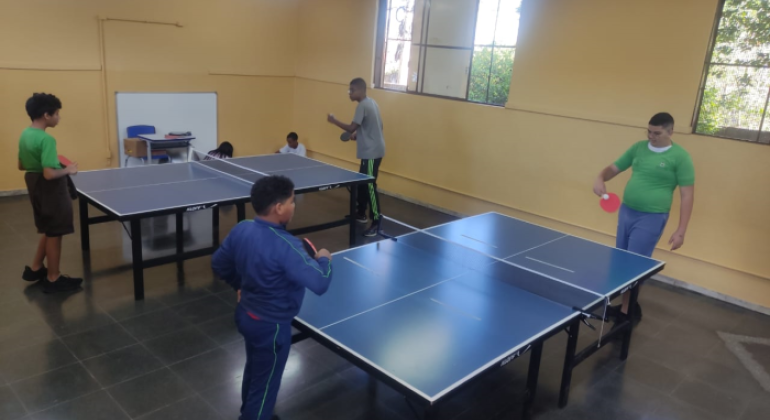 #paratodosverem: a foto mostra uma sala, com paredes amarelas e amplas janelas gradeadas. No centro da sala, há duas mesas de tênis de mesa onde quatro alunos estão envolvidos na atividade esportiva, com raquetes nas mãos e atentos ao jogo. As crianças vestem uniformes escolares ou roupas esportivas. Ao fundo, duas crianças estão sentadas próximo à parede.