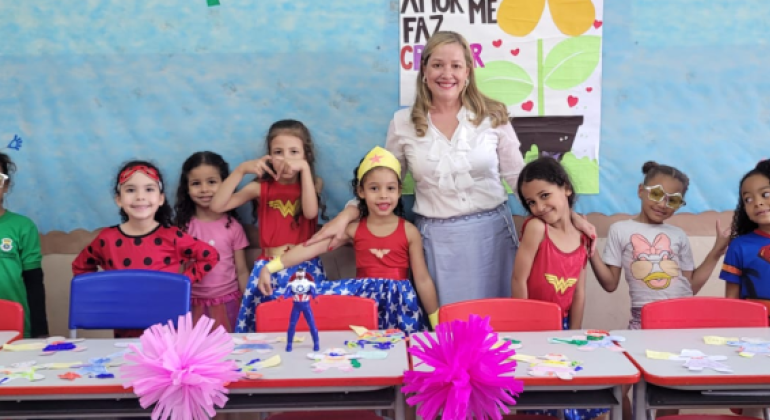 #paratodosverem: na foto, oito meninas fantasiadas de super-heroínas e uma professora posam sorridentes atrás de mesas com trabalhos manuais coloridos, em uma sala de aula. Atrás da professora, há um quadro decorado de azul claro e moldura de papel marrom.