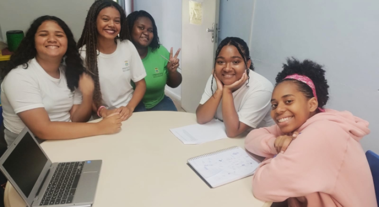 #paratodosverem: na imagem, cinco estudantes estão sentadas em torno de uma mesa redonda, sorrindo para a câmera. Três usam camisetas brancas com o brasão da Prefeitura de Belo Horizonte; outra está com camisa de uniforme verde contendo o brasão, e a quinta veste moletom rosa. Sobre a mesa, há um notebook e cadernos abertos. Ao fundo, há uma parede azul-clara, uma porta branca e um cartaz com a palavra “PAS”.