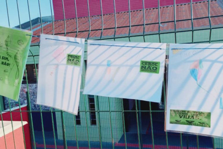 #paratodosverem: Cartazes em papel verde estão presos a uma grade metálica. No centro, há o desenho de uma pipa simples, contornada em preto, com a linha esticada em direção ao canto inferior. Ao redor, aparecem pequenos desenhos de nuvens. Em destaque, a frase em letras grandes e pretas: “PIPA SIM, CEROL NÃO!”.