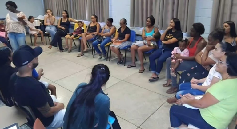 #paratodosverem: em uma sala de aula, há pessoas organizadas em círculo, sentadas em cadeiras, criando um ambiente de diálogo e acolhimento. Ao centro da cena, uma mulher em pé segura papéis. O grupo é formado majoritariamente por mulheres, algumas com crianças no colo ou ao seu lado. A sala tem paredes claras, janelas com cortinas e ventiladores de parede.