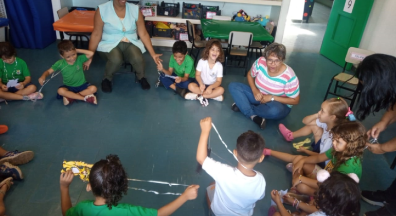 #paratodosverem: na imagem, tirada em uma perspectiva de cima para baixo, diversas crianças uniformizadas estão sentadas em círculo, no chão de uma sala. Entre elas, há três mulheres adultas. Atrás delas, estão três prateleiras brancas cheias de brinquedos, algumas cadeiras e duas mesas pequenas (uma com toalha alaranjada e outra com toalha verde). No canto superior direito, há uma porta aberta com o número nove estampado.