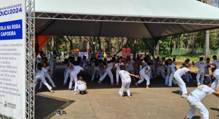 #paratodosverem: foto de uma apresentação de capoeira ao ar livre, com dezenas de crianças uniformizadas em roda, jogando em duplas sob uma tenda. À esquerda, um painel destaca a importância da capoeira como patrimônio cultural e instrumento de luta contra o racismo. O evento integra a 3ª Semana da Educação BHEDUCA 2024.