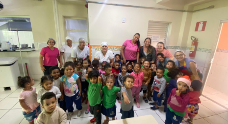 Alimentação saudável na prática #paratodosverem: Um grupo de crianças pequenas posa sorridente em uma sala com piso branco e paredes claras. Atrás delas estão várias mulheres adultas, algumas com toucas e uniformes de cozinha. Ao fundo há uma faixa parcialmente visível com a palavra “ALIMENTAÇÃO” e um extintor de incêndio fixado na parede.